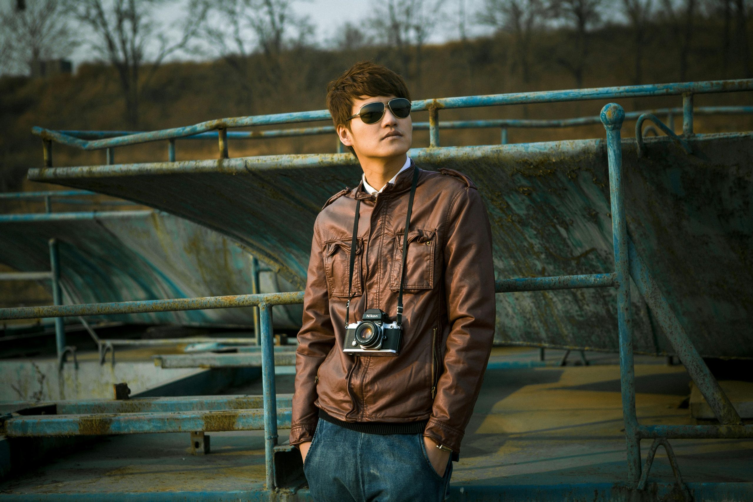 Stylish man wearing sunglasses and leather jacket holding a vintage camera in an outdoor setting.