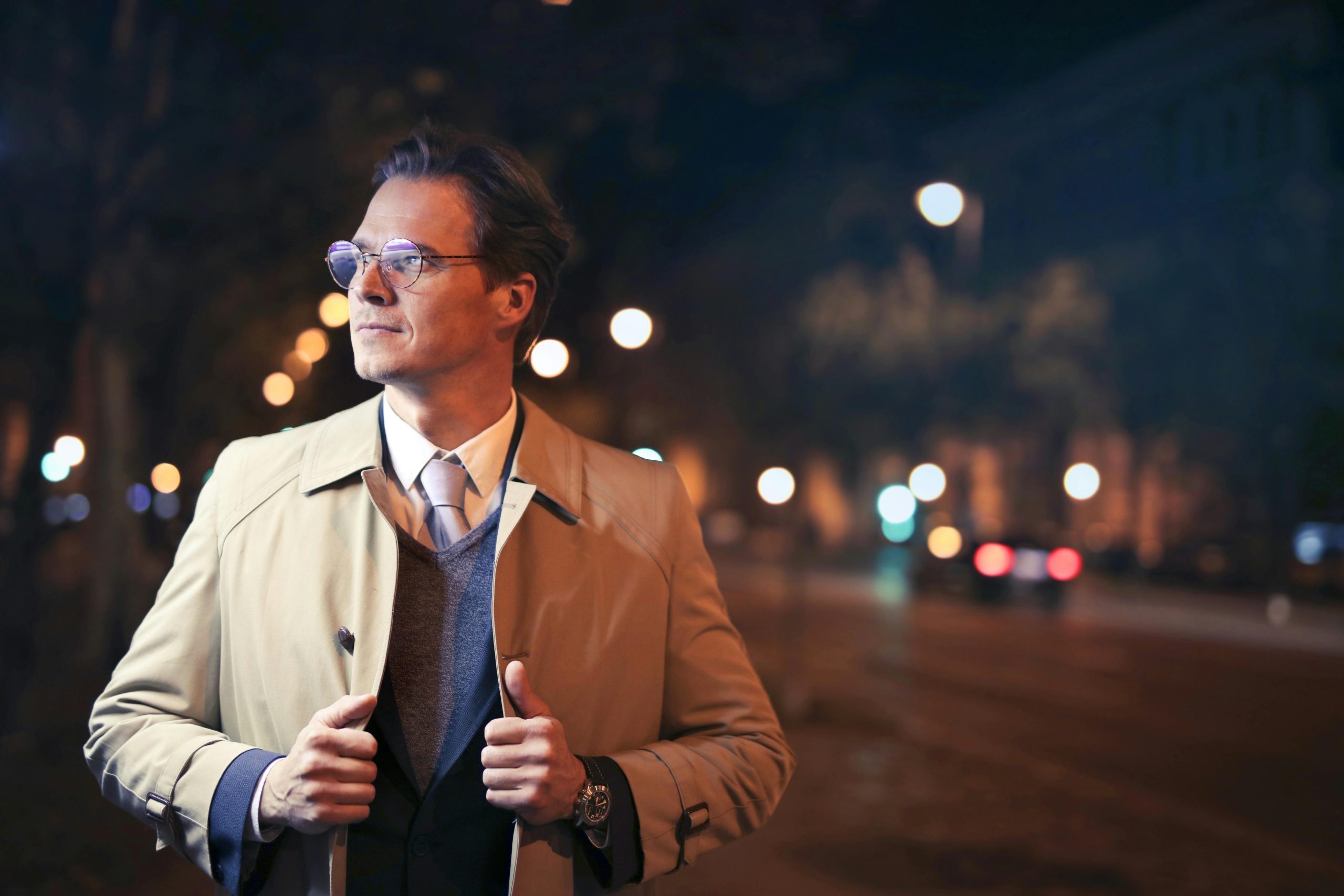 Elegant man in a trench coat walking city streets at night with bokeh lights.