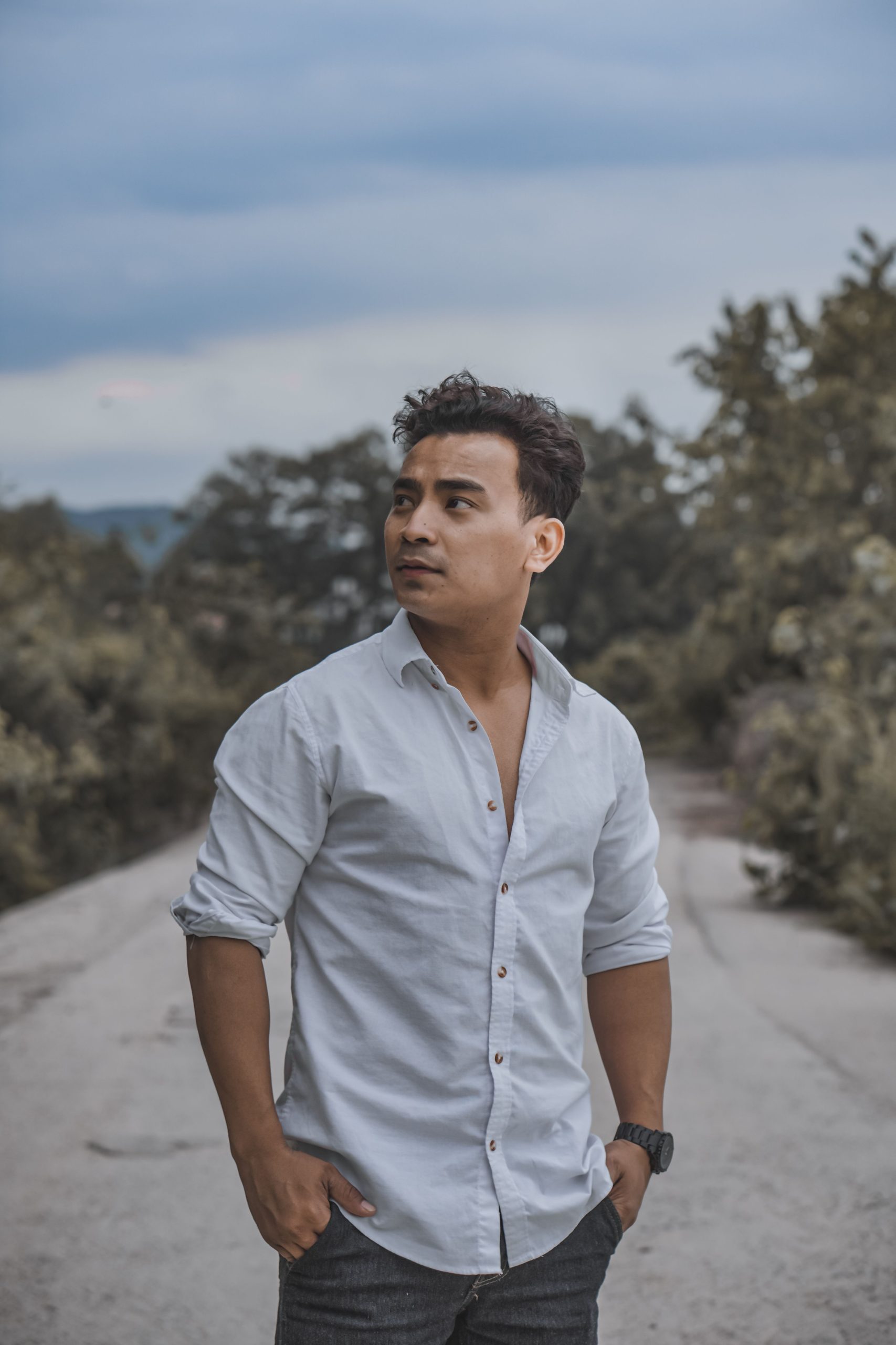 A man in casual white shirt and jeans posing confidently on a rural road with trees around.
